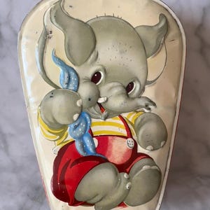 Könnte beinhalten: Vintage Metallbox mit einem Cartoon-Elefanten in roter Latzhose und einem gelb-weiß gestreiften Hemd. Der Elefant hat graue Haut, große Ohren und eine blaue Schleife. Die Dose hat einen cremefarbenen Hintergrund.