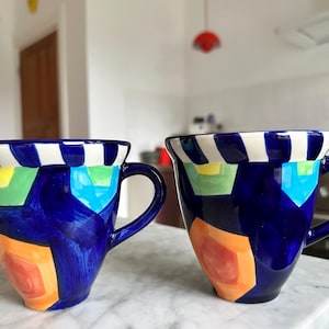 Può includere: Due tazze in ceramica con un design astratto e vibrante. Le tazze sono principalmente blu scuro con forme geometriche colorate in arancione, verde e giallo. I bordi presentano strisce bianche e nere e ogni tazza ha un manico curvo.