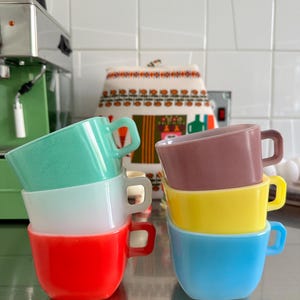 Könnte beinhalten: Zwei Stapel kleiner, quadratischer Tassen mit Henkeln. Der linke Stapel besteht aus einer grünen Tasse oben, einer weißen in der Mitte und einer roten unten. Der rechte Stapel besteht aus einer malvenfarbenen Tasse oben, einer gelben in der Mitte und einer blauen unten.