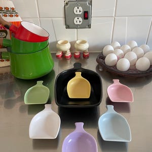 Könnte beinhalten: Ein Set aus sieben bunten Keramikschaufeln in verschiedenen Farbtönen von Grün, Gelb, Rosa, Weiß, Hellblau und Lila. Die Schaufeln sind in Form einer kleinen, schmalen Schüssel mit einem Griff. Die Schaufeln sind auf einer silbernen Oberfläche angeordnet.