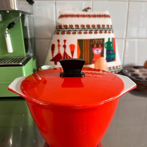 Può includere: Una pirofila rosso brillante con coperchio abbinato e pomello nero. La ciotola presenta bordi interni e manici laterali bianchi, con una finitura liscia e lucida. Il recipiente ha una forma conica e arrotondata ed è centrato nell'inquadratura.