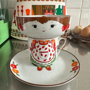 Può includere: Un set vintage di ciotola, tazza e piattino in ceramica. La ciotola ha la forma di un viso di ragazza con capelli castani e un fiocco rosso. La tazza e il piattino presentano un design a fragole e decorazioni floreali. Il set è su una superficie in acciaio inossidabile.
