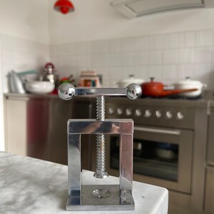 May include: A silver nutcracker with a screw mechanism and two round handles. The nutcracker is sitting on a white marble countertop.