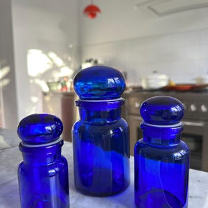 Puede incluir: Tres botes de farmacia de vidrio azul cobalto con tapas redondeadas. Los botes son de diferentes tamaños y se asientan sobre una superficie de mármol. Los botes tienen un diseño clásico y probablemente se utilizan para almacenamiento o exhibición.