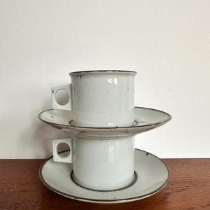Puede incluir: Dos tazas de café de cerámica blanca apiladas con platillos a juego. Las tazas tienen asas cuadradas y un borde oscuro. Los platillos tienen un borde oscuro a juego. Las tazas y los platillos están sobre una superficie de madera.