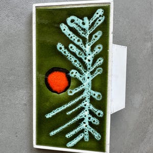 May include: A rectangular, mid-century modern wall tile with a white metal frame. The tile features a dark green background with a light blue, stylized tree design and a red circle. The tile is mounted on a white metal bracket.