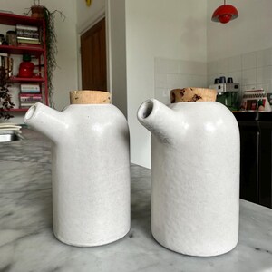 May include: Two cylindrical off-white ceramic oil dispensers with a matte finish, each featuring a side-angled spout and a natural light brown cork stopper. The vessels have a smooth, rounded top design and a sturdy base for upright placement.
