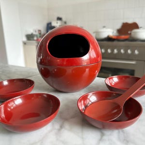 May include: A red lacquered serving set comprising a large spherical container with an oval opening, four small bowls, and a serving spoon. The set is displayed on a marble countertop in a kitchen. The bowls and spoon are oval-shaped.