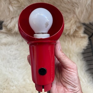 May include: A red, vintage lamp with a white lightbulb. The lamp features a rounded shade and a cylindrical base with a black button. The lightbulb has text printed on it. The lamp is held in a hand.