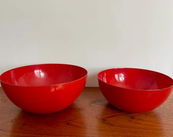 May include: Two glossy red bowls of different sizes sit on a wooden surface. The bowls have a smooth, rounded shape and a vibrant, solid red colour. The bowls are empty and ready to be used.