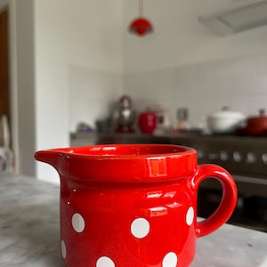 May include: A red ceramic jug with white polka dots. The jug has a handle and a spout. The kitchenware is on a marble countertop. The background is a blurred kitchen scene.