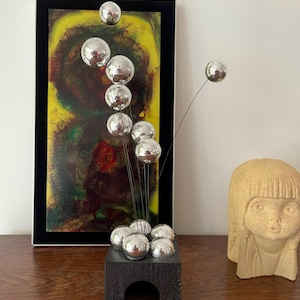 Pode incluir: Uma escultura moderna com várias esferas prateadas presas a finos fios pretos, emergindo de um cubo de madeira escura. A escultura é exibida sobre uma superfície de madeira, com uma pintura abstrata emoldurada ao fundo.