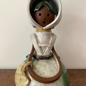 May include: A ceramic figurine of a woman wearing a white bonnet and a green dress. She has brown skin and black hair. She is holding a brown strap in her hand.