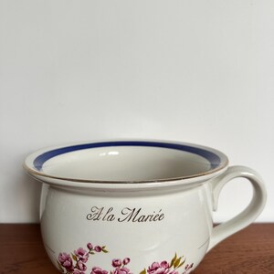 Può includere: Vaso da notte in ceramica bianca con bordo blu e finiture dorate. Il vaso presenta un motivo floreale con fiori rosa e foglie verdi. Le parole "À la Mariée" sono scritte in corsivo nero.
