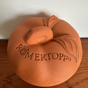 Peut inclure: Un couvercle en terre cuite de couleur terracotta pour un récipient de cuisson, en forme de pomme stylisée avec une tige centrale et une feuille. Le mot RÖMERTOPF est gravé en lettres capitales sur la surface supérieure de la céramique.