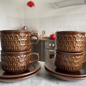 Puede incluir: Dos juegos de tazas de té y platillos de cerámica marrón. Cada juego tiene dos tazas apiladas una encima de la otra. Las tazas tienen un patrón geométrico marrón y dorado.