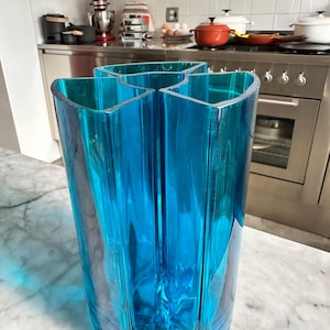 May include: A tall, turquoise-colored glass vase with a unique, multi-sectioned design. The vase has a geometric shape with four distinct sections that come together at the base. The transparent glass allows light to pass through, creating a play of light and shadow.