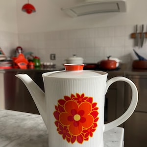 以下が含まれることがあります： 赤、オレンジ、黄色の花柄が描かれた白いセラミック製のコーヒーポット。ポットには注ぎ口とハンドルが付いています。