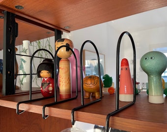 May include: A wooden shelf displays a collection of decorative items. Included are Japanese Kokeshi dolls, a ceramic dog figurine, and other colorful figurines. The shelf is made of wood and has black metal dividers to hold the items in place.