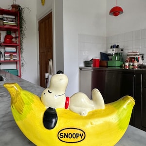 Könnte beinhalten: Eine Keramikfigur von Snoopy, die auf einer gelben Banane liegt. Die Banane hat grüne und gelbe Farbverläufe, mit einem schwarzen Oval und dem Wort "SNOOPY" in einem weißen Oval. Snoopy ist weiß mit einem schwarzen Fleck und einem roten Halsband.
