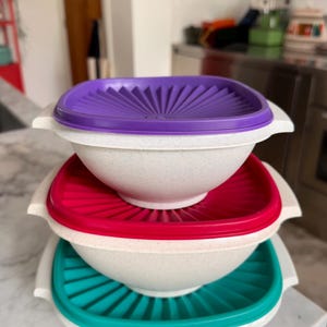 May include: A stack of three off-white speckled bowls with matching lids. The top lid is purple, the middle lid is magenta, and the bottom lid is teal. Each lid features a sunburst pattern of radiating ridges and side handles for gripping.