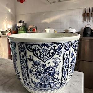 May include: A large, round ceramic planter with a white and blue floral design. The planter has a wide rim and a ribbed base. The blue floral pattern is intricate and covers the entire surface. The planter is on a marble surface.