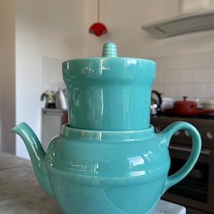 Op de afbeelding: Een tealblauwe keramische theepot met een bijpassend deksel en een ingebouwd filter. De theepot heeft een rond lichaam en een tuit aan de zijkant.