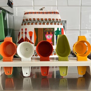 Op de afbeelding: Een set kleurrijke meetinstrumenten in oranje, wit, rood, groen en geel, weergegeven op een wit rek. De instrumenten omvatten maatbekers en -lepels, perfect om te bakken en te koken. Een keukenhanddoek met patroon is op de achtergrond.