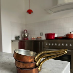 May include: A set of three copper saucepans with brass handles. The saucepans are stacked on top of each other, with the smallest saucepan on top. The saucepans are in focus, while the background is blurred.