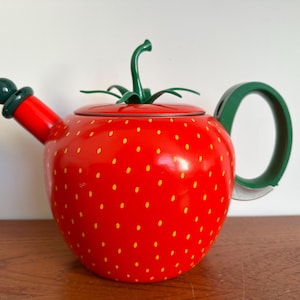 Könnte beinhalten: Eine rote Teekanne in Erdbeerform mit gelben Punkten, einem grünen Griff und einem grünen Stiel. Der Ausguss und der Deckel sind ebenfalls grün. Die Teekanne steht auf einer Holzoberfläche.