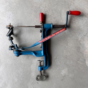May include: A blue metal PEFRA Kartoffel-u.Obst-Schälmaschine potato and fruit peeler featuring a hand crank with a red handle, a vertical adjustment knob with a red handle, a metal blade, and a screw-style clamp base for mounting to a surface.