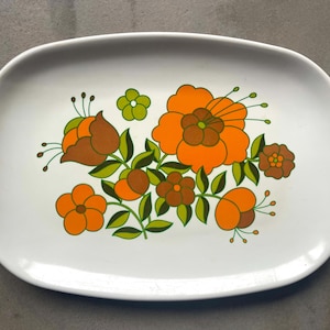 Può includere: Piatto da portata ovale bianco con un motivo floreale in tonalità arancio, marrone e verde. I fiori sono stilizzati e hanno un aspetto retrò.