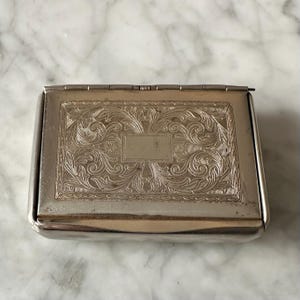 Pode incluir: Uma caixa retangular de metal prateado com tampa articulada. A tampa e a frente da caixa apresentam um design floral gravado e ornamentado. A caixa está fechada e assenta numa superfície de mármore.