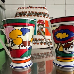 May include: Two decorative ceramic mugs with a landscape design. The mugs have white handles and bases with blue stripes. The artwork features houses, trees, and clouds in bright colours, including red, yellow, and blue. Each mug is approximately 10 cm tall.
