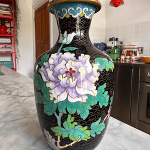 Pode incluir: Um vaso cloisonné preto com uma flor de peônia roxa e branca em destaque e folhas verdes. A superfície é decorada com um padrão de fio de ouro fino sobre um fundo preto, com uma borda e base em tom dourado.