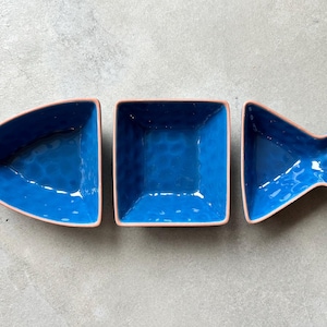 Op de afbeelding: Drie keramische serveerschalen in de vorm van een vis. De schalen zijn helderblauw met een getextureerd oppervlak en een lichtroze rand. De schalen zijn gerangschikt in de vorm van een vis.