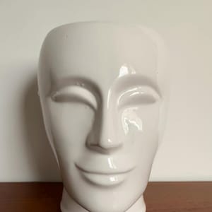 Può includere: Un vaso bianco in ceramica a forma di volto con gli occhi chiusi e un leggero sorriso. Il vaso ha una finitura liscia e lucida e una base arrotondata. Il design è minimalista e moderno, adatto all'arredamento della casa.