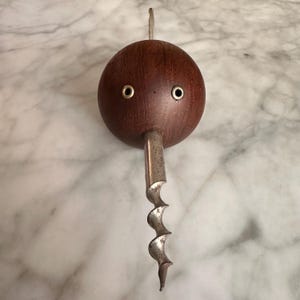May include: A spherical dark brown wooden corkscrew featuring two small circular metal eyelets on the front surface and a silver-toned metal spiral worm extending from the bottom, with a thin metal handle protruding from the top of the sphere.