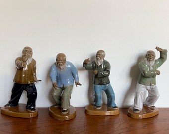 May include: Four ceramic figurines of elderly individuals with long white beards, each posed in a different tai chi movement. They wear jackets in brown, light blue, dark green, and sage green, paired with black, tan, blue, or white trousers.