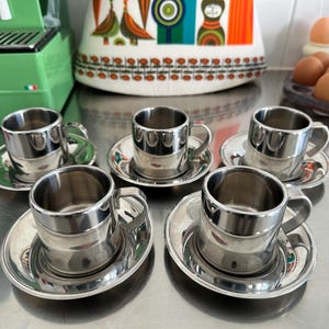 Könnte beinhalten: Set aus sechs Espresso-Tassen und Untertassen aus Edelstahl. Die Tassen haben ein klassisches Design mit Henkel und stehen auf passenden runden Untertassen. Das Set wird auf einer reflektierenden Oberfläche präsentiert.