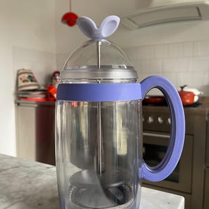 Könnte beinhalten: Eine transparente Glas-French-Press-Kaffeemaschine mit einem lila Deckel und Griff. Der Deckel hat zwei kleine, lila, hasenohr-förmige Griffe.