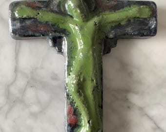 May include: A ceramic wall cross featuring a glossy, light green figure of a person centered on a dark, mottled background with shades of black, blue, and red. The cross has a smooth, reflective finish and a small circular hole at the top for hanging.