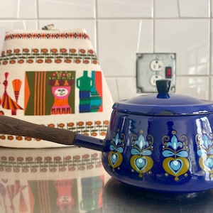 Op de afbeelding: Een blauwe emaille pan met een houten handvat en een bloemenpatroon. De pan heeft een deksel en is versierd met blauwe, gele en groene bloemen.