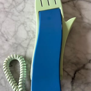 May include: A vintage telephone with a unique hand-shaped design. The phone features a light green hand-shaped receiver and a blue center section. The coiled cord and base are also light green. The phone rests on a marble surface.