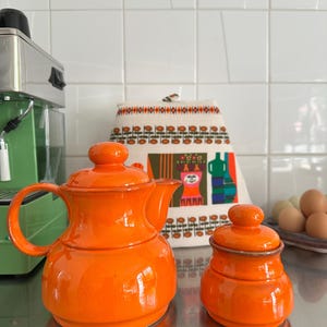 Könnte beinhalten: Helles orangefarbenes Keramik-Teeservice mit Teekanne und Zuckerdose. Die Teekanne hat einen gebogenen Griff und einen Ausguss. Die Zuckerdose hat einen Deckel mit einem kleinen Knopf. Das Set steht auf einer Edelstahl-Oberfläche. Im Hintergrund eine grüne Espressomaschine.