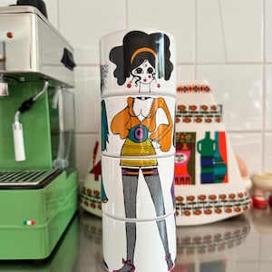 Puede incluir: Una pila de cuatro tazas de cerámica blanca con una ilustración retro de una mujer con un atuendo de estilo mod. La mujer tiene cabello negro, ojos grandes y lleva una blusa naranja, una falda amarilla y medias negras. Las tazas están apiladas una encima de la otra.