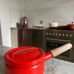 Puede incluir: Una cacerola de esmalte rojo brillante con un mango de madera y una tapa. La cacerola está sobre una encimera de mármol blanco.