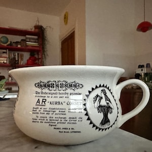 Op de afbeelding: Crèmekleurige keramische mok met een grote handgreep, met zwarte tekst en illustraties. De tekst bevat "AR 'KURBA' CORSET" en een belofte van omruiling. Een cirkelvormig embleem toont een vrouw in een korset.