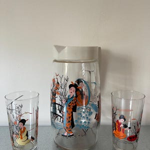 以下が含まれることがあります： 日本のデザインが施されたガラス製のピッチャーとタンブラー3点セット。ピッチャーとタンブラーは、白い背景に、伝統的な日本の服装をした女性たちのカラフルなイラストが描かれています。