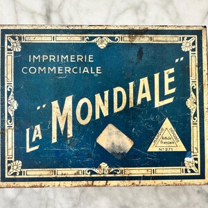 May include: Vintage rectangular metal box with a distressed blue and cream design. The box features the text "IMPRIMERIE COMMERCIALE" and "LA MONDIALE" in white lettering. The box has a decorative border and a small triangle logo.